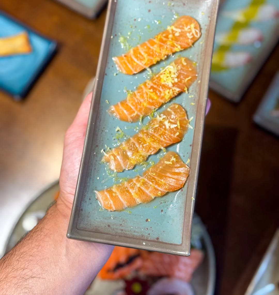 Carpaccio de salmão maçaricado num prato azul segurado sobre a mesa