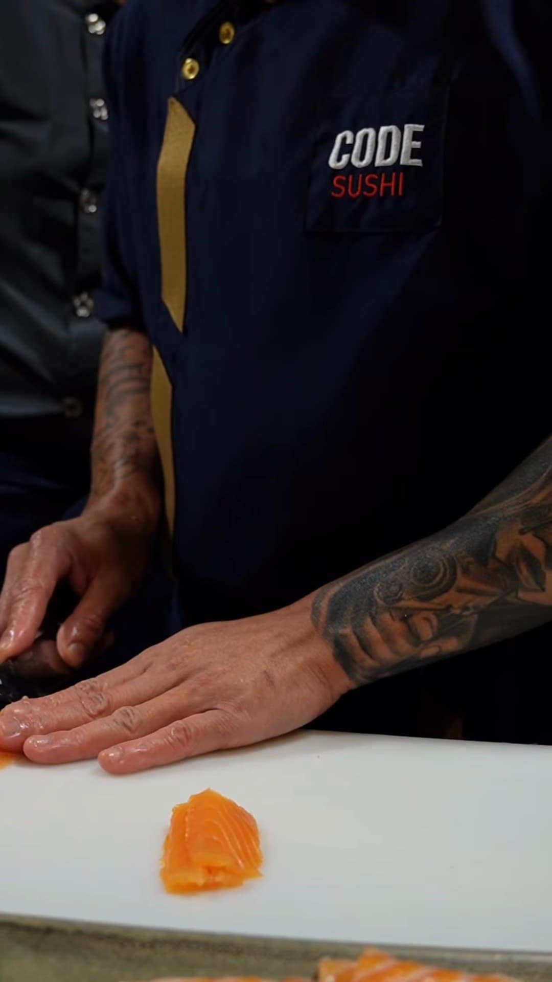 Mãos tatuadas cortando peixe na bancada, com o dólmã do Code Sushi