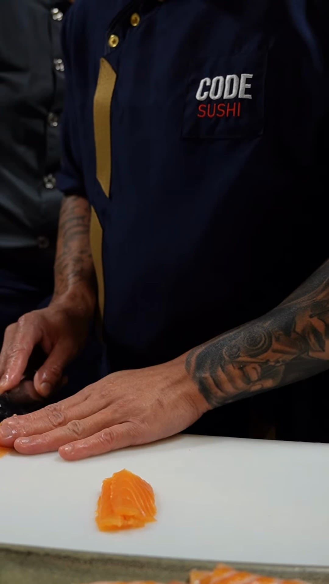 Mãos tatuadas cortando peixe na bancada, com o dólmã do Code Sushi