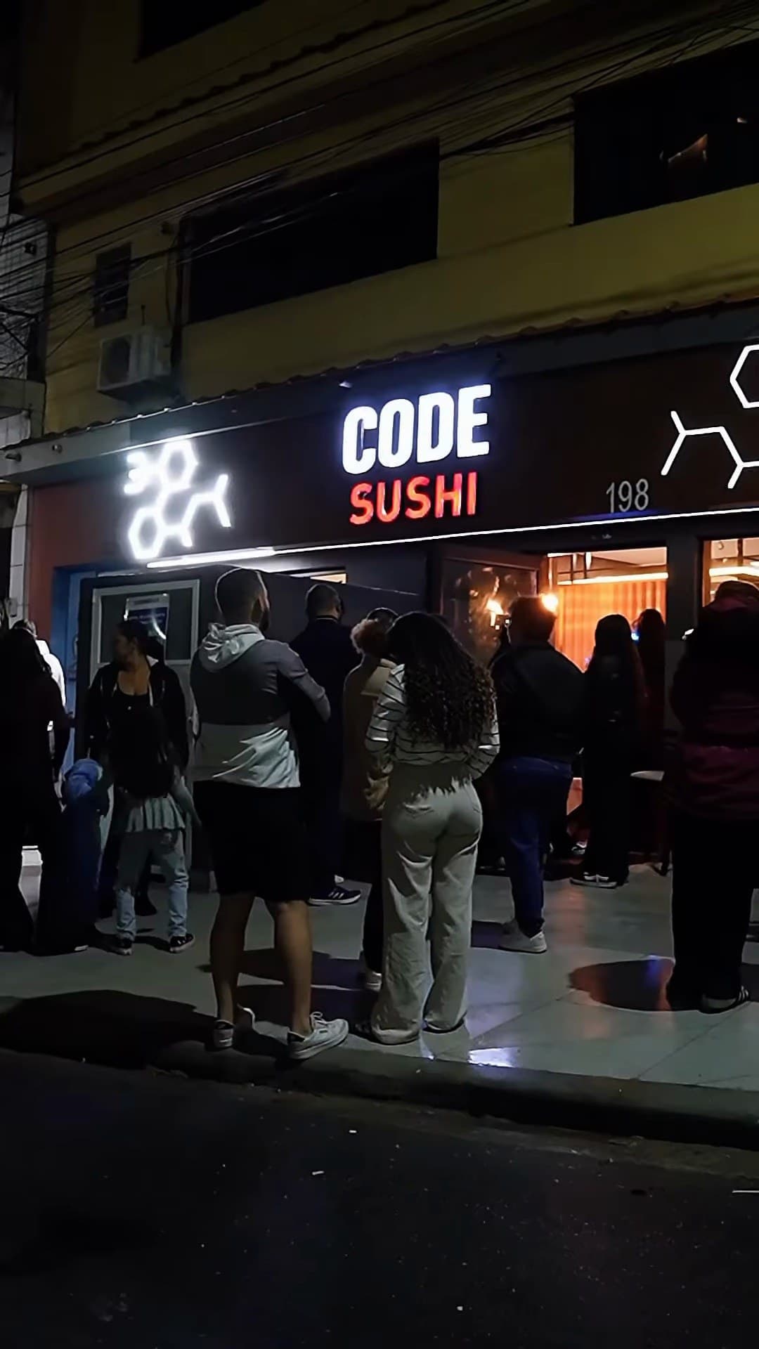 A fachada do Code Sushi à noite, com o letreiro aceso e pessoas na calçada