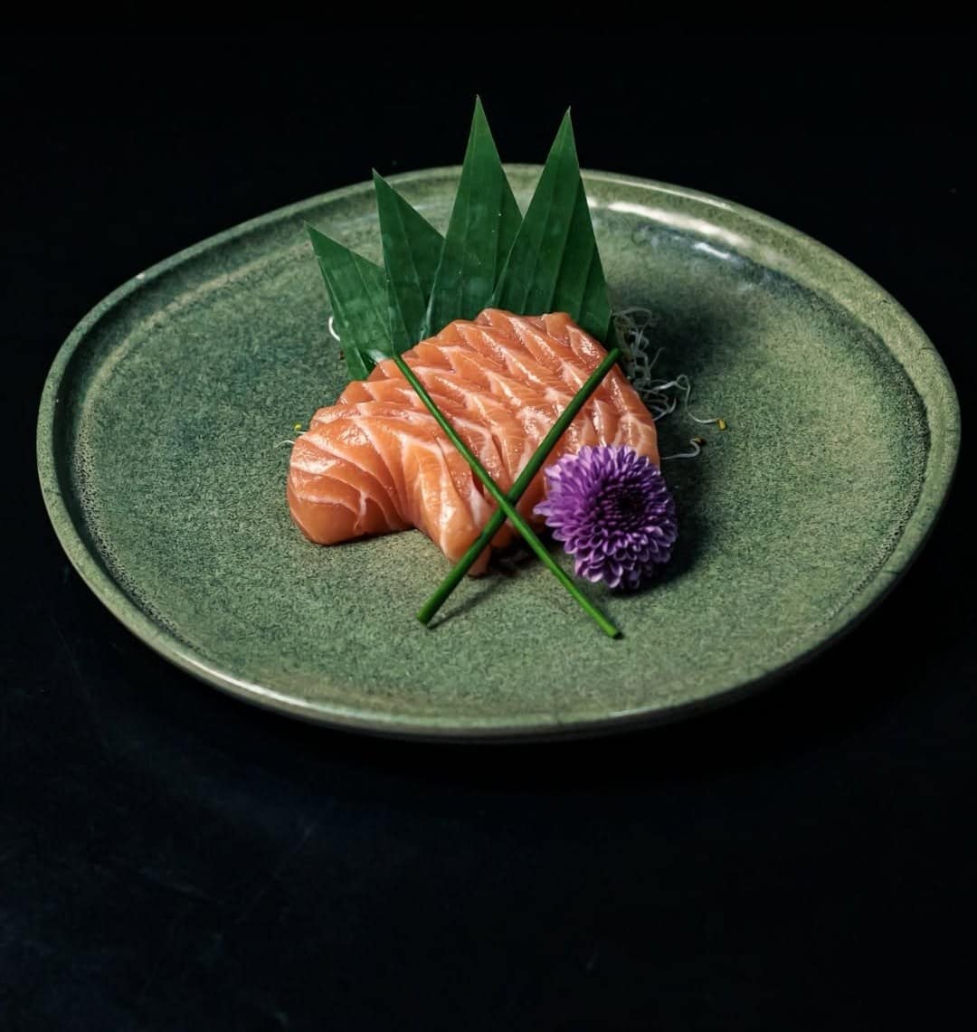 Sashimi de salmão com folhas e uma flor roxa num prato verde, sobre fundo preto