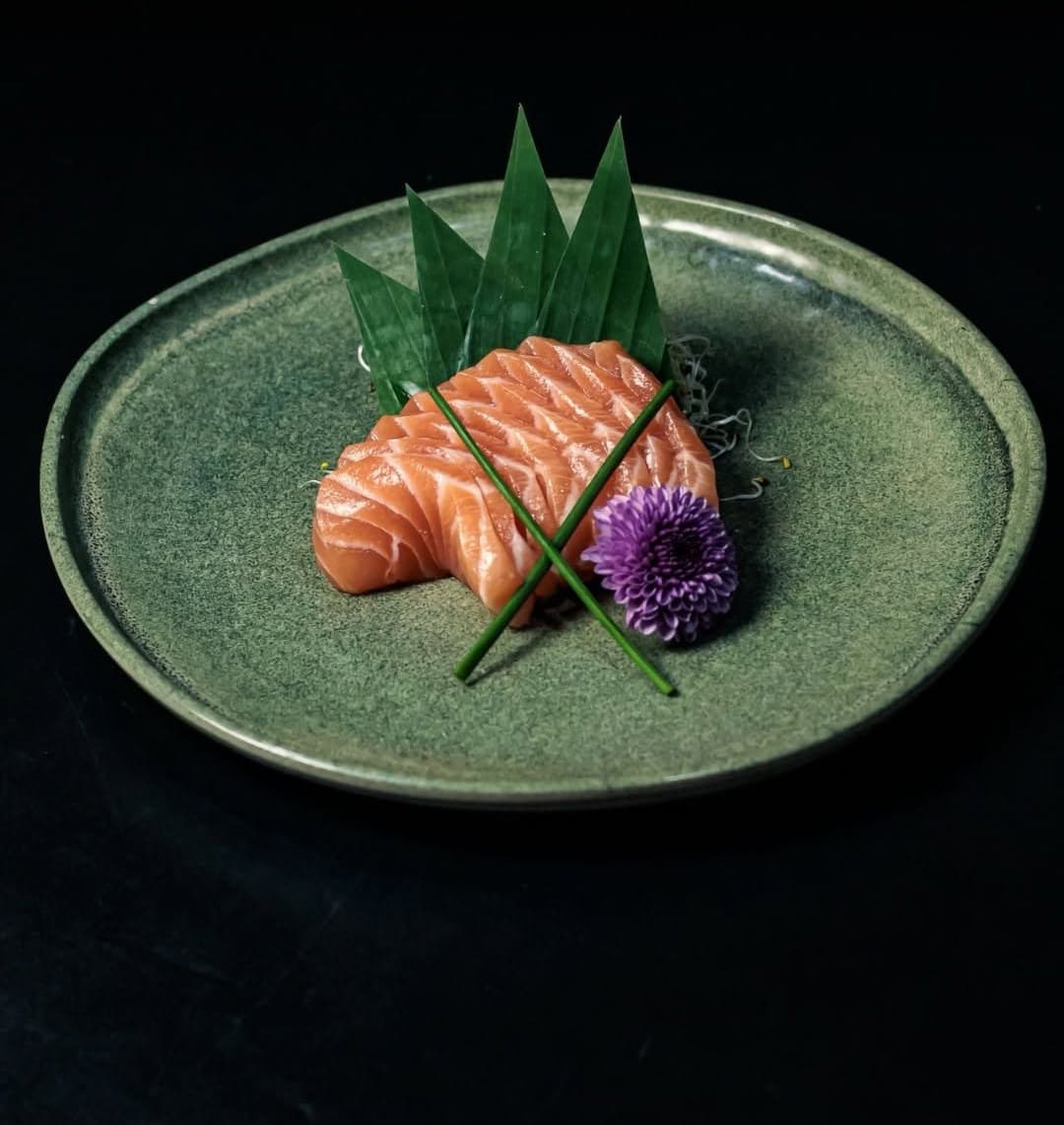 Sashimi de salmão com folhas e uma flor roxa num prato verde, sobre fundo preto